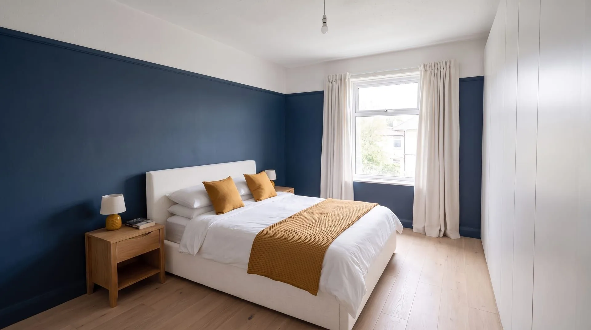 The same bedroom with deep navy walls, an off-white headboard and curtains, and mustard cushions, throw and lamp base