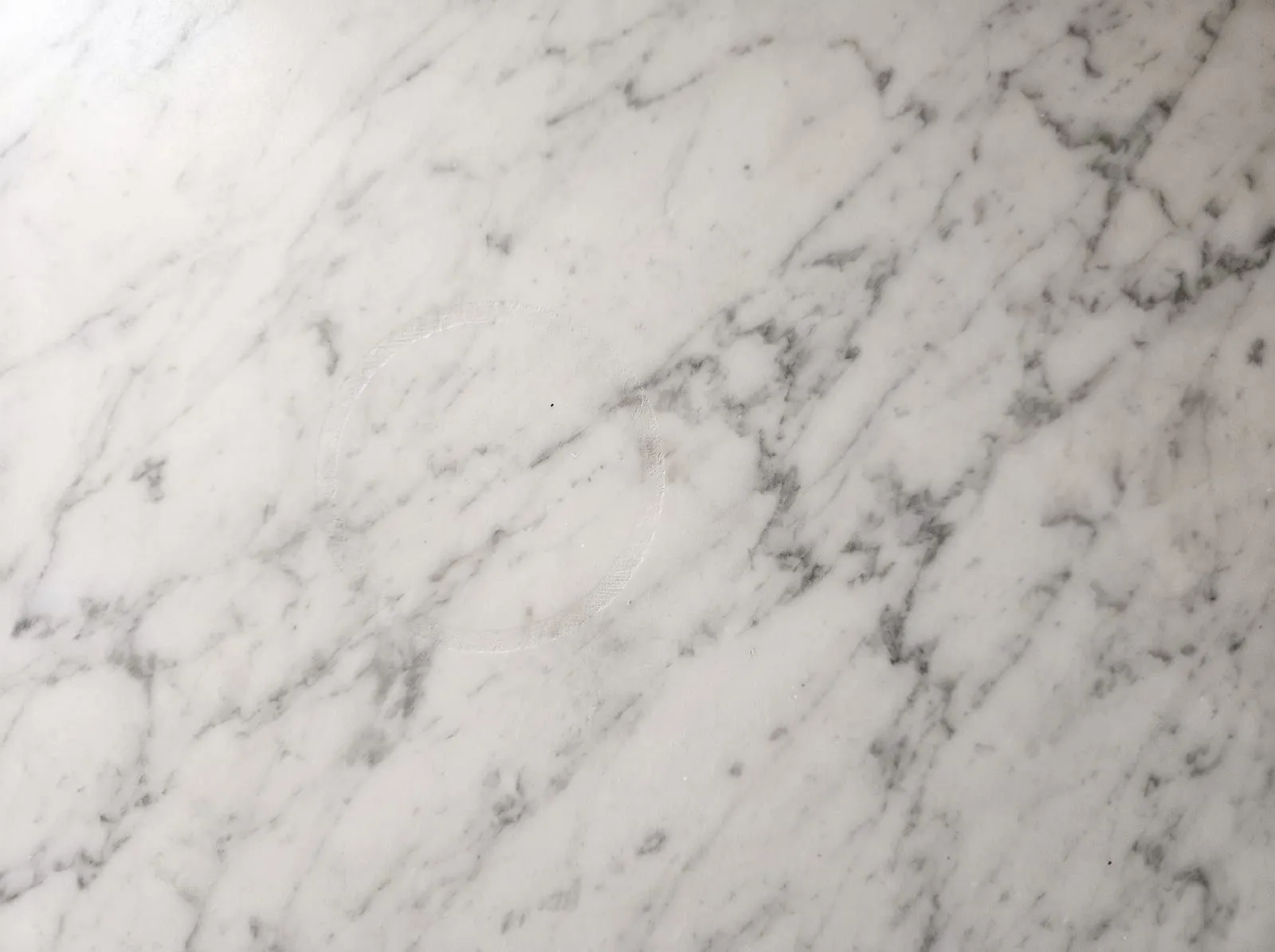 A close-up of honed white marble with grey veins and a faint dull etch ring left by an acidic drink