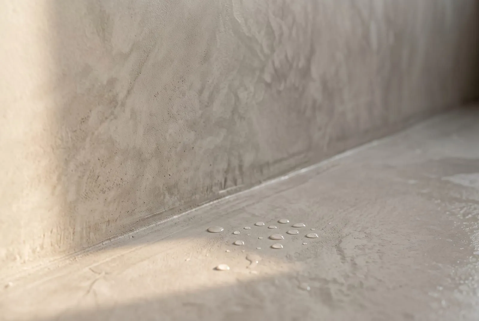A close-up of sealed microcement where a wall meets a shower floor, with water beading on the surface