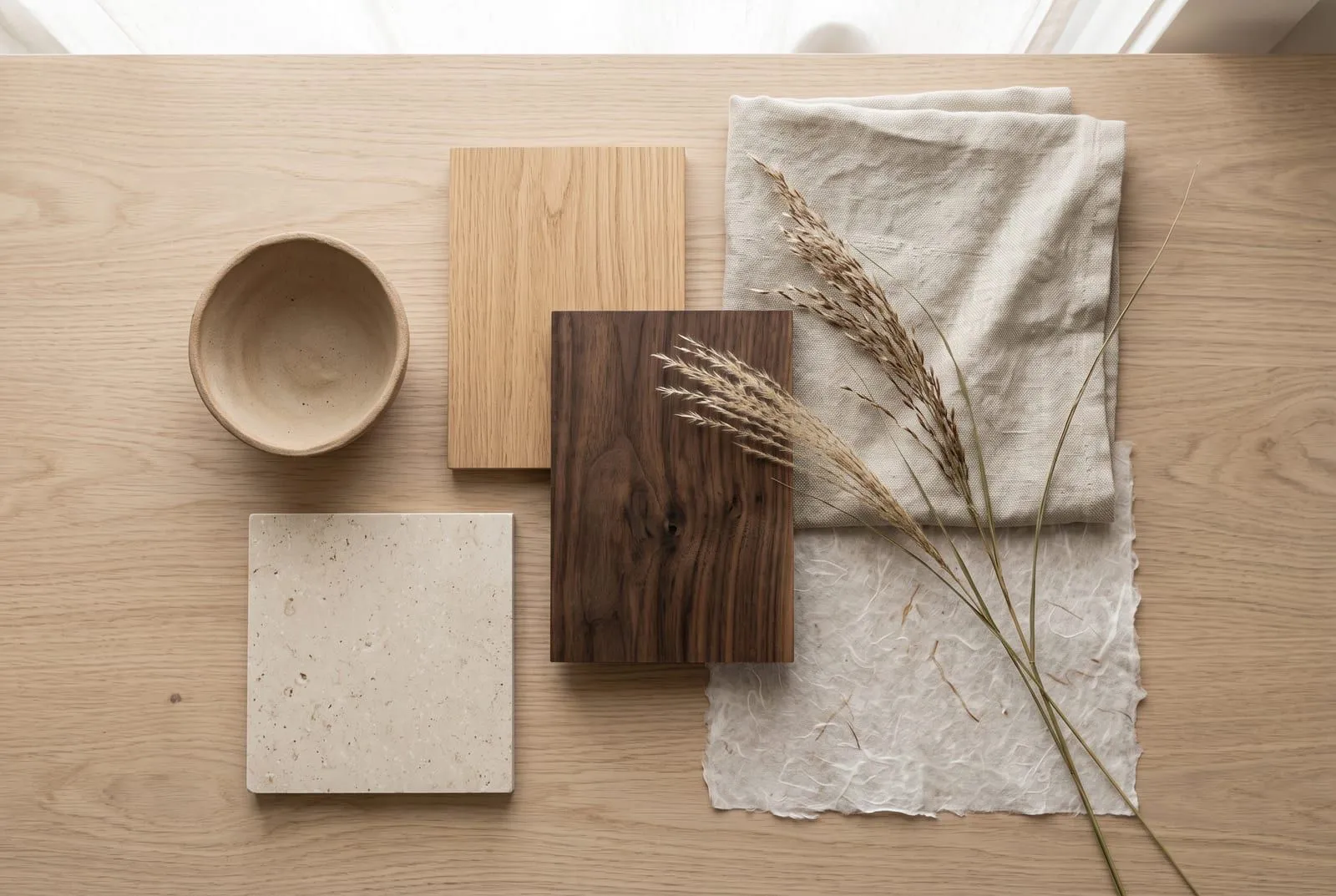 A flat lay of Japandi materials: light oak, dark walnut, raw linen, rice paper, a matte clay bowl, a stone tile and a sprig of dried grass