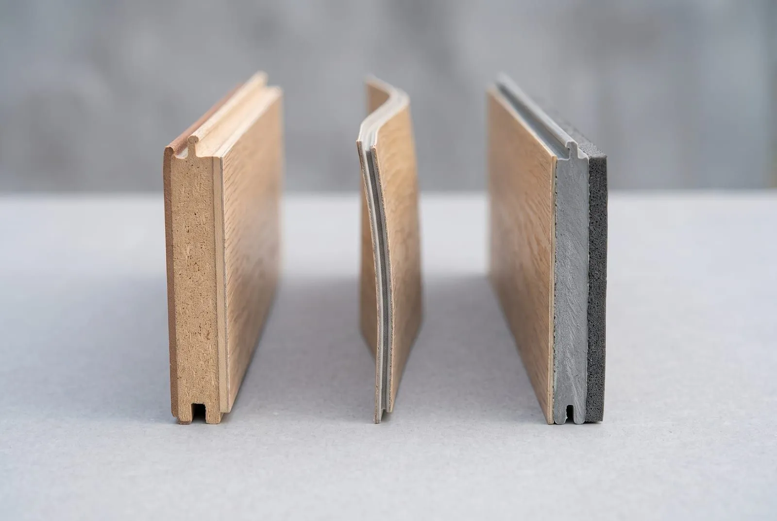 Three flooring planks cut open side by side: laminate with a thick fibreboard core, a thin flexible vinyl plank, and a rigid grey SPC core with a foam pad underneath