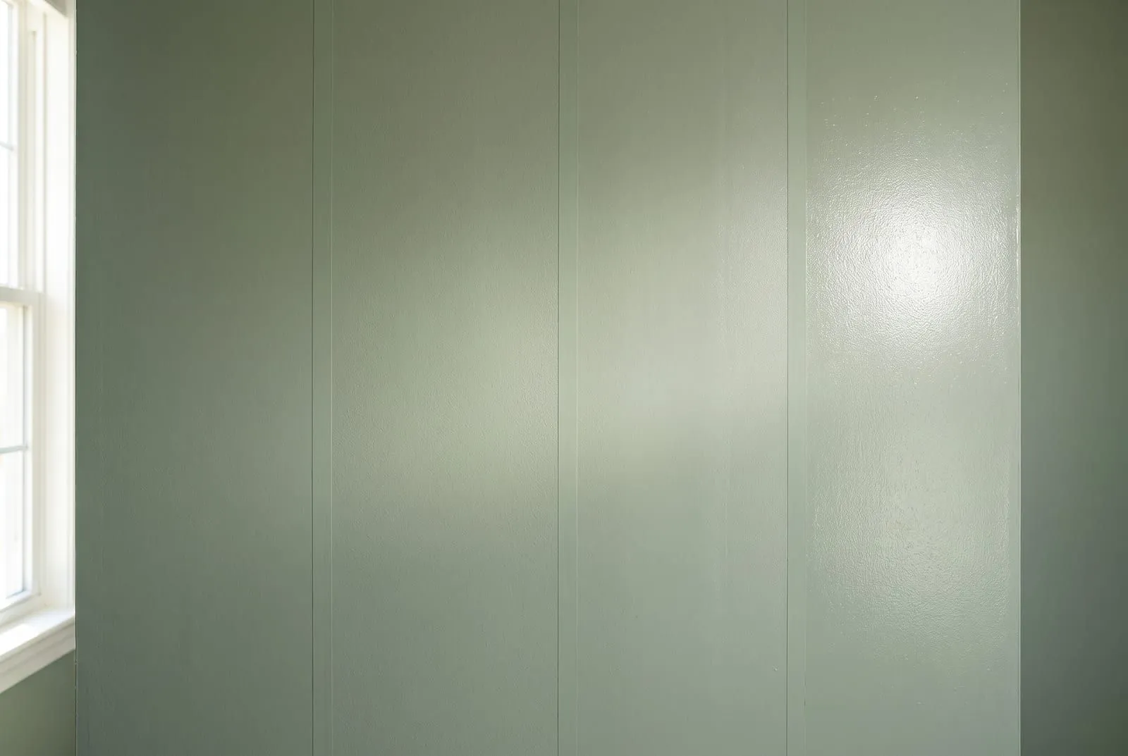 Close-up of a sage green wall painted in four vertical bands from flat to semi-gloss, under low window light